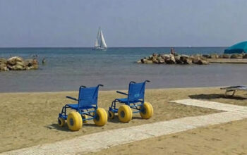 Due sedie JOB azzurre per la balneazione dei disabili posizionate sulla sabbia accanto a una passerella bianca, su una spiaggia attrezzata con ombrelloni e una barca a vela sullo sfondo.