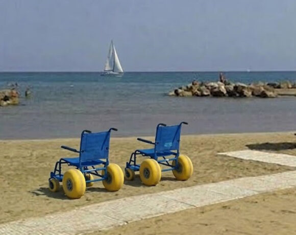 Due sedie JOB azzurre per la balneazione dei disabili posizionate sulla sabbia accanto a una passerella bianca, su una spiaggia attrezzata con ombrelloni e una barca a vela sullo sfondo.