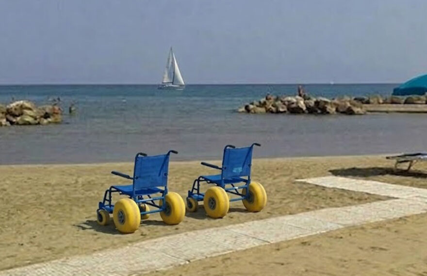 Due sedie JOB azzurre per la balneazione dei disabili posizionate sulla sabbia accanto a una passerella bianca, su una spiaggia attrezzata con ombrelloni e una barca a vela sullo sfondo.