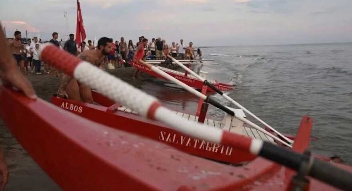 Pattini di salvataggio rossi pronti sulla battigia dell'Albos Club di Fregene per il Palio della Voga con bagnini e pubblico sullo sfondo.