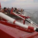 Pattini di salvataggio rossi pronti sulla battigia dell'Albos Club di Fregene per il Palio della Voga con bagnini e pubblico sullo sfondo.