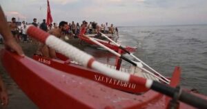 Pattini di salvataggio rossi pronti sulla battigia dell'Albos Club di Fregene per il Palio della Voga con bagnini e pubblico sullo sfondo.