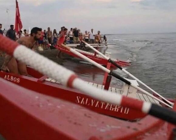 Pattini di salvataggio rossi pronti sulla battigia dell'Albos Club di Fregene per il Palio della Voga con bagnini e pubblico sullo sfondo.