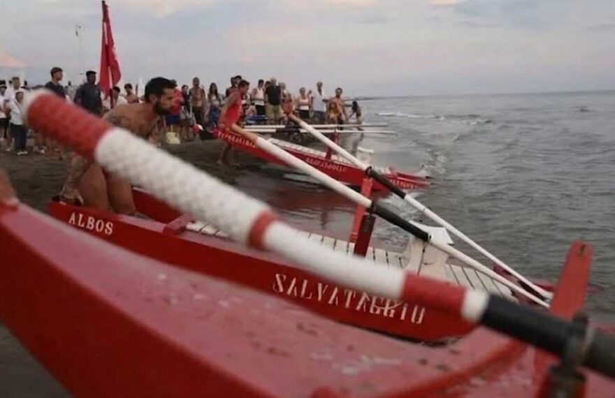 Pattini di salvataggio rossi pronti sulla battigia dell'Albos Club di Fregene per il Palio della Voga con bagnini e pubblico sullo sfondo.