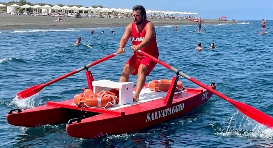 Bagnino rema su un pattino di salvataggio rosso in mare, con ciambella di salvataggio a bordo.