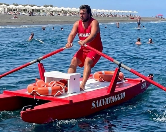 Bagnino rema su un pattino di salvataggio rosso in mare, con ciambella di salvataggio a bordo.