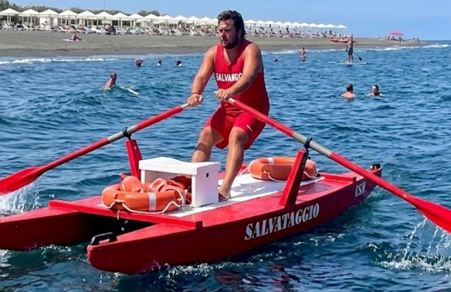 Bagnino rema su un pattino di salvataggio rosso in mare, con ciambella di salvataggio a bordo.