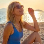 donna in costume da bagno con occhiali da sole seduta sulla spiaggia al tramonto