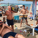 Una scena balneare caotica e affollata in Italia, che illustra vari comportamenti fastidiosi dei bagnanti citati nel decalogo del bon ton del Codacons, come l'uso di telefoni, musica ad alto volume, fumo e rumore.