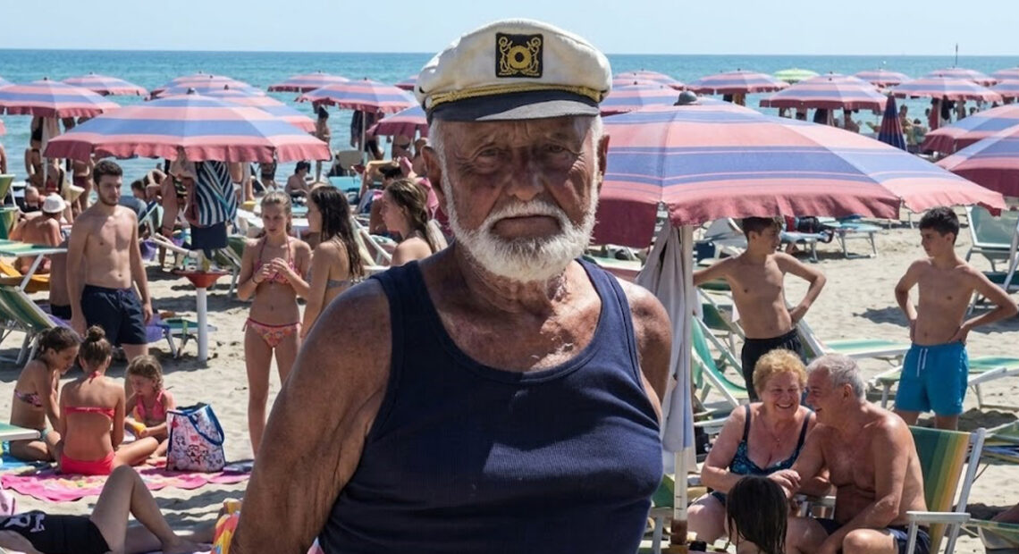 Primo piano del bagnino Guerrino Ricci con il cappello da capitano sulla sua spiaggia di Riccione, circondato da clienti sotto gli ombrelloni.