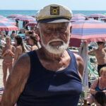 Primo piano del bagnino Guerrino Ricci con il cappello da capitano sulla sua spiaggia di Riccione, circondato da clienti sotto gli ombrelloni.