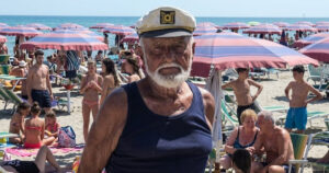 Primo piano del bagnino Guerrino Ricci con il cappello da capitano sulla sua spiaggia di Riccione, circondato da clienti sotto gli ombrelloni.