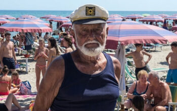 Primo piano del bagnino Guerrino Ricci con il cappello da capitano sulla sua spiaggia di Riccione, circondato da clienti sotto gli ombrelloni.