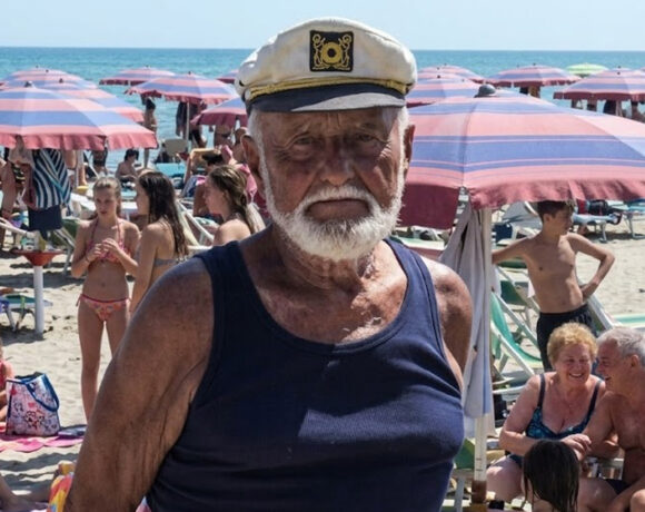 Primo piano del bagnino Guerrino Ricci con il cappello da capitano sulla sua spiaggia di Riccione, circondato da clienti sotto gli ombrelloni.