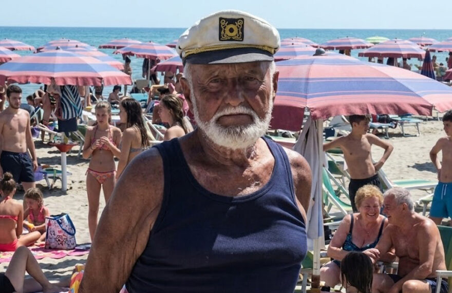 Primo piano del bagnino Guerrino Ricci con il cappello da capitano sulla sua spiaggia di Riccione, circondato da clienti sotto gli ombrelloni.