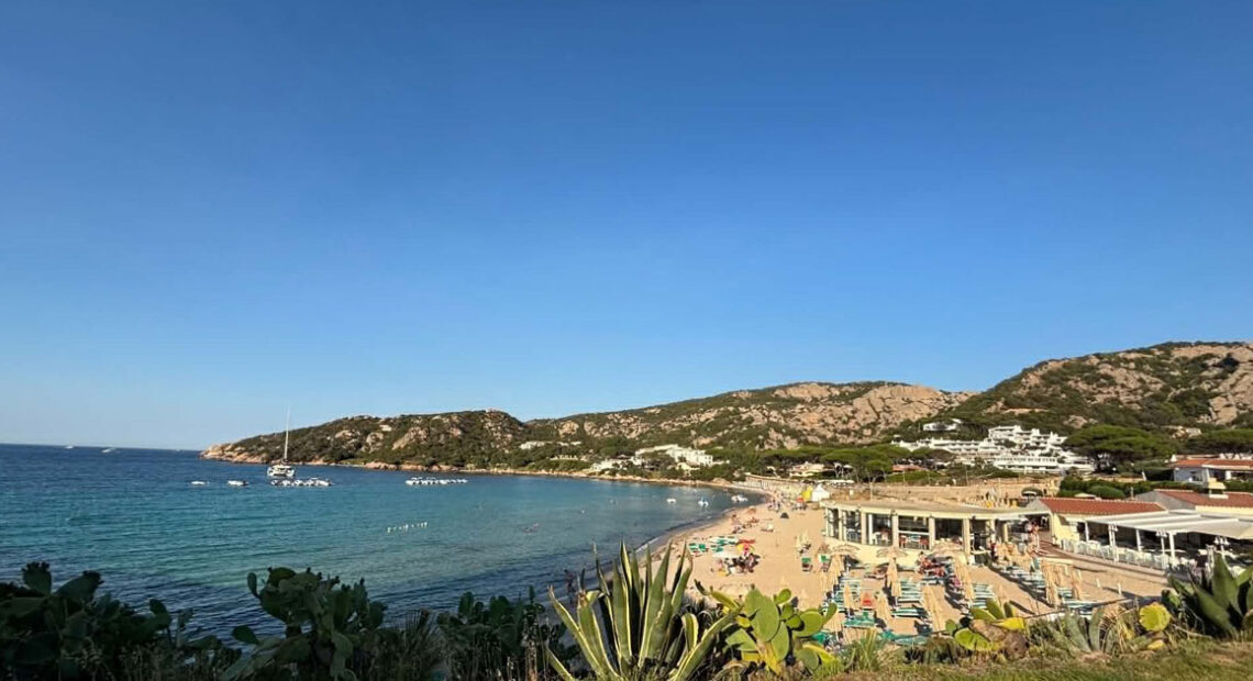Vista panoramica della spiaggia di Mannena ad Arzachena, con acque cristalline, bagnanti e colline rocciose della Costa Smeralda sotto un cielo sereno.