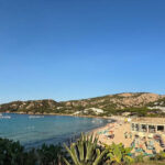 Vista panoramica della spiaggia di Mannena ad Arzachena, con acque cristalline, bagnanti e colline rocciose della Costa Smeralda sotto un cielo sereno.