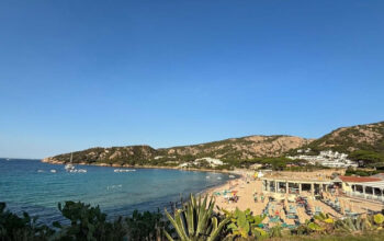 Vista panoramica della spiaggia di Mannena ad Arzachena, con acque cristalline, bagnanti e colline rocciose della Costa Smeralda sotto un cielo sereno.