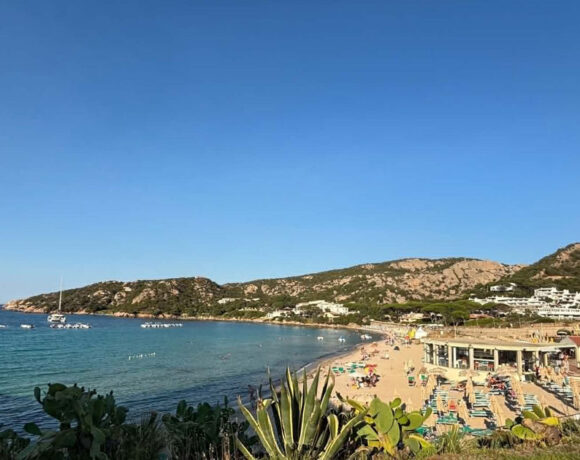 Vista panoramica della spiaggia di Mannena ad Arzachena, con acque cristalline, bagnanti e colline rocciose della Costa Smeralda sotto un cielo sereno.