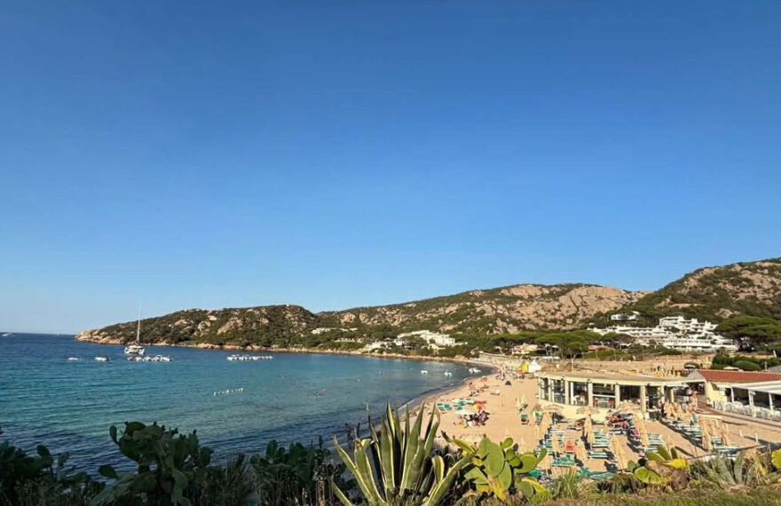 Vista panoramica della spiaggia di Mannena ad Arzachena, con acque cristalline, bagnanti e colline rocciose della Costa Smeralda sotto un cielo sereno.
