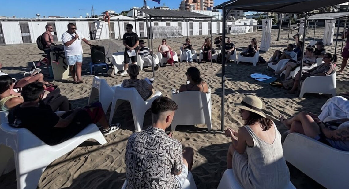 Un gruppo di ragazzi e relatori under 35 seduti in cerchio sulla spiaggia durante un incontro culturale organizzato da Informagiovani.
