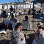 Un gruppo di ragazzi e relatori under 35 seduti in cerchio sulla spiaggia durante un incontro culturale organizzato da Informagiovani.
