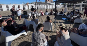 Un gruppo di ragazzi e relatori under 35 seduti in cerchio sulla spiaggia durante un incontro culturale organizzato da Informagiovani.