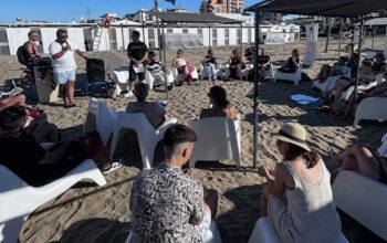 Un gruppo di ragazzi e relatori under 35 seduti in cerchio sulla spiaggia durante un incontro culturale organizzato da Informagiovani.