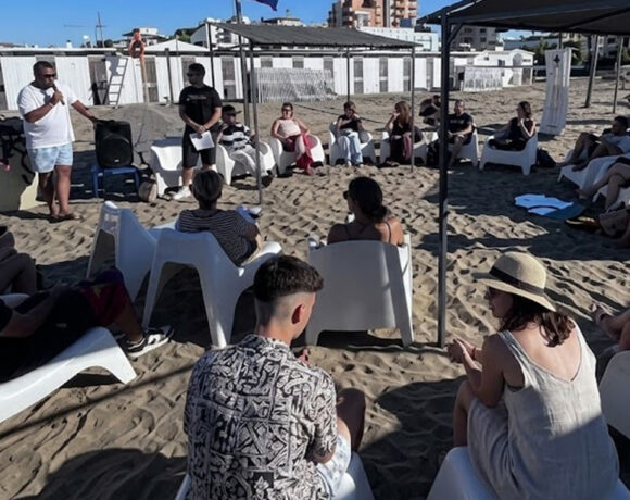 Un gruppo di ragazzi e relatori under 35 seduti in cerchio sulla spiaggia durante un incontro culturale organizzato da Informagiovani.