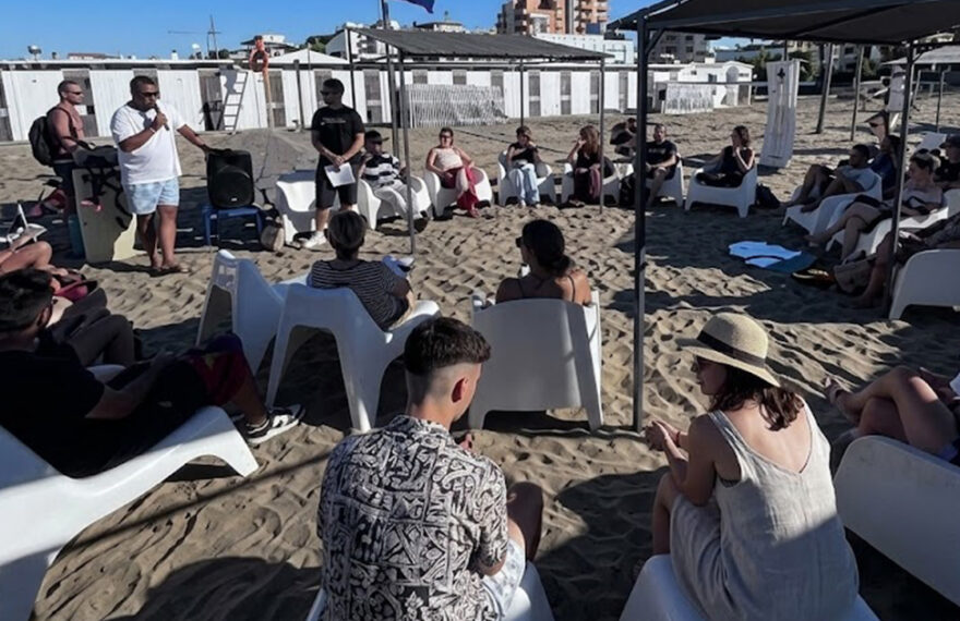 Un gruppo di ragazzi e relatori under 35 seduti in cerchio sulla spiaggia durante un incontro culturale organizzato da Informagiovani.