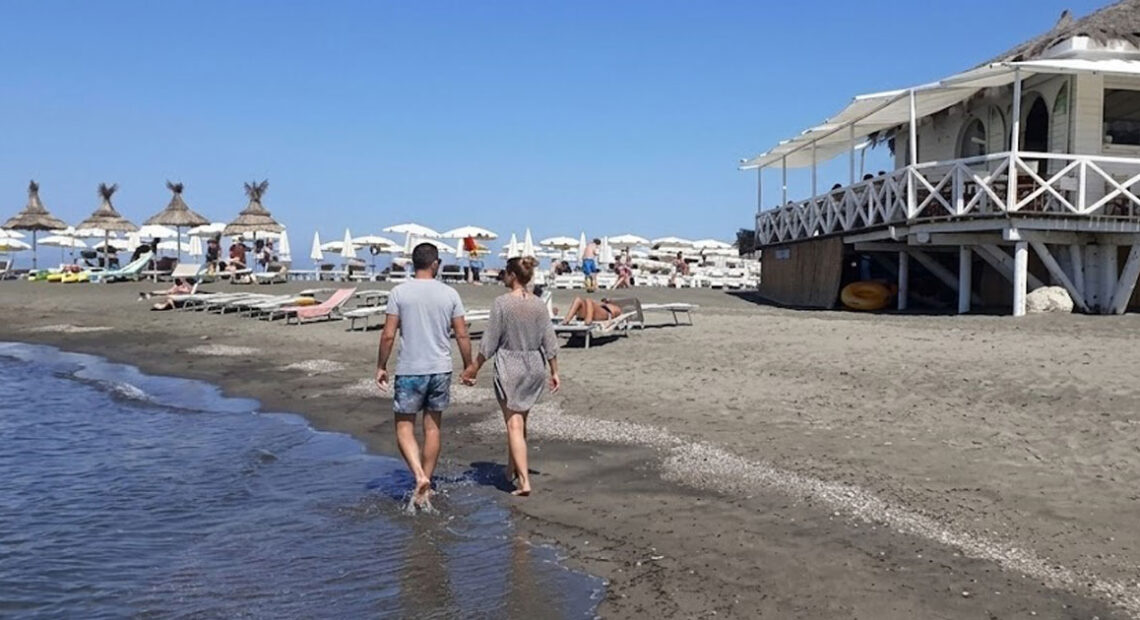 Una coppia cammina sul bagnasciuga della spiaggia di Ladispoli con stabilimento balneare in legno sullo sfondo