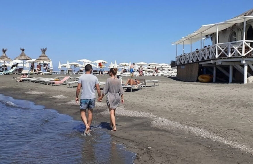 Una coppia cammina sul bagnasciuga della spiaggia di Ladispoli con stabilimento balneare in legno sullo sfondo