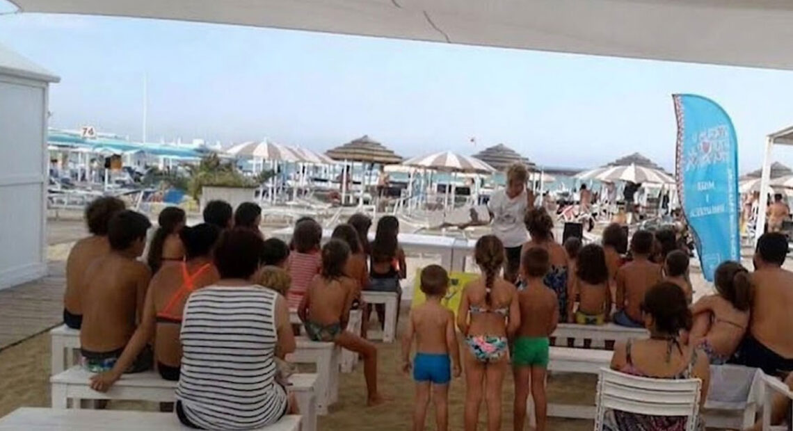 Un gruppo di bambini e genitori seduti su panchine bianche all'ombra in uno stabilimento balneare di Riccione, mentre assistono a una lettura animata in spiaggia.