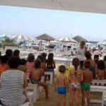 Un gruppo di bambini e genitori seduti su panchine bianche all'ombra in uno stabilimento balneare di Riccione, mentre assistono a una lettura animata in spiaggia.