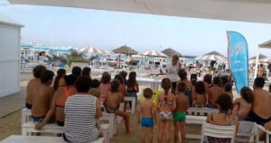 Un gruppo di bambini e genitori seduti su panchine bianche all'ombra in uno stabilimento balneare di Riccione, mentre assistono a una lettura animata in spiaggia.
