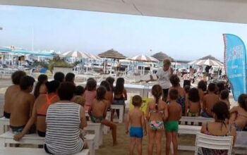 Un gruppo di bambini e genitori seduti su panchine bianche all'ombra in uno stabilimento balneare di Riccione, mentre assistono a una lettura animata in spiaggia.