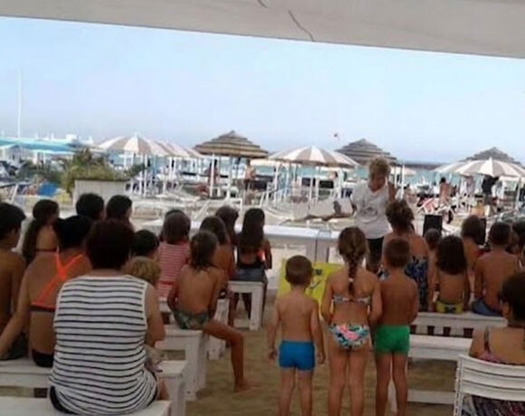 Un gruppo di bambini e genitori seduti su panchine bianche all'ombra in uno stabilimento balneare di Riccione, mentre assistono a una lettura animata in spiaggia.
