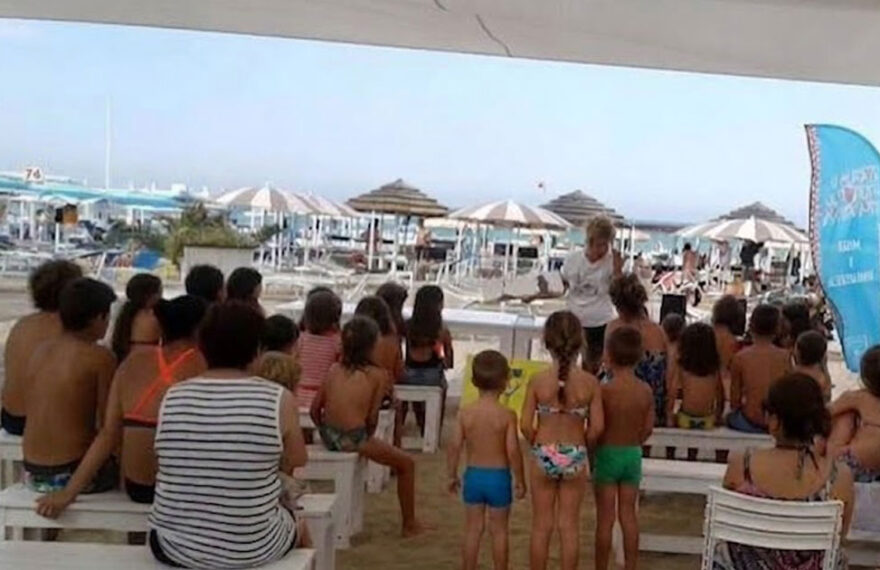 Un gruppo di bambini e genitori seduti su panchine bianche all'ombra in uno stabilimento balneare di Riccione, mentre assistono a una lettura animata in spiaggia.