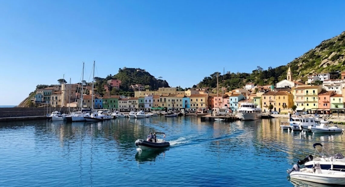 Vista panoramica del molo e delle case colorate di Giglio Porto con barche ormeggiate e acqua cristallina.