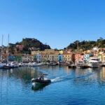 Vista panoramica del molo e delle case colorate di Giglio Porto con barche ormeggiate e acqua cristallina.
