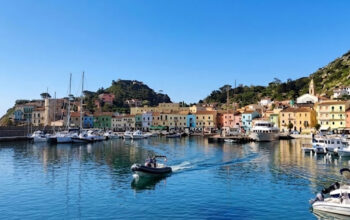 Vista panoramica del molo e delle case colorate di Giglio Porto con barche ormeggiate e acqua cristallina.