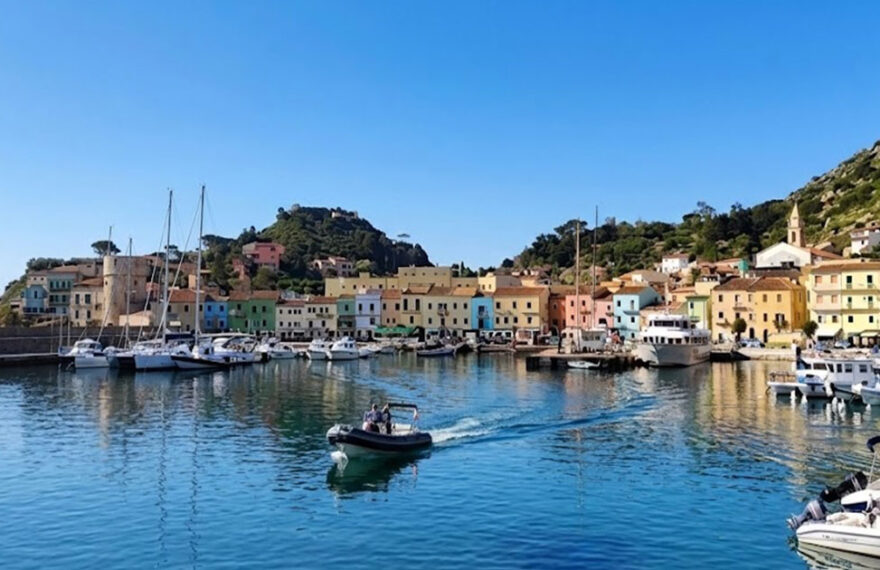 Vista panoramica del molo e delle case colorate di Giglio Porto con barche ormeggiate e acqua cristallina.
