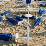 Spiaggia devastata dai maranza al Bagno Holiday di Lido di Spina, con ombrelloni blu sradicati e lettini rovesciati sulla sabbia