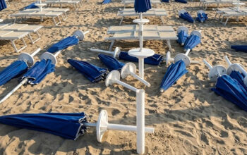 Spiaggia devastata dai maranza al Bagno Holiday di Lido di Spina, con ombrelloni blu sradicati e lettini rovesciati sulla sabbia
