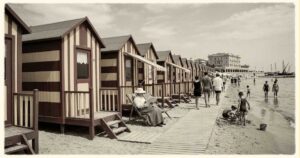 Ricostruzione d'epoca di una fila di cabine balneari in legno a righe sulla spiaggia con bagnanti nei primi del Novecento.