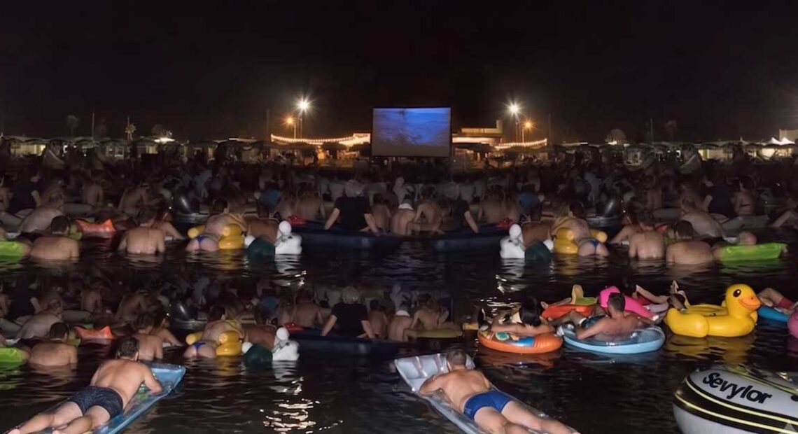Spettatori in mare su materassini e gonfiabili mentre guardano il film Lo Squalo sul bagnasciuga di Fano.