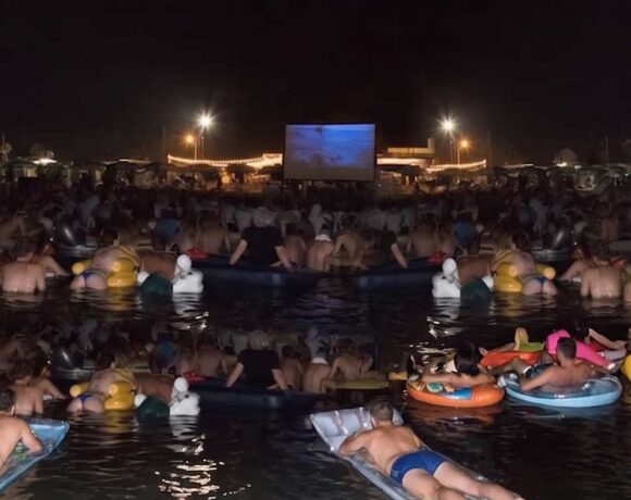 Spettatori in mare su materassini e gonfiabili mentre guardano il film Lo Squalo sul bagnasciuga di Fano.