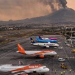 Vista panoramica dell'aeroporto di Catania con aerei di EasyJet e Ryanair a terra e una densa nube di fumo vulcanico che si alza dall'Etna sullo sfondo al tramonto.