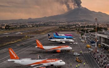 Vista panoramica dell'aeroporto di Catania con aerei di EasyJet e Ryanair a terra e una densa nube di fumo vulcanico che si alza dall'Etna sullo sfondo al tramonto.