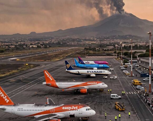 Vista panoramica dell'aeroporto di Catania con aerei di EasyJet e Ryanair a terra e una densa nube di fumo vulcanico che si alza dall'Etna sullo sfondo al tramonto.